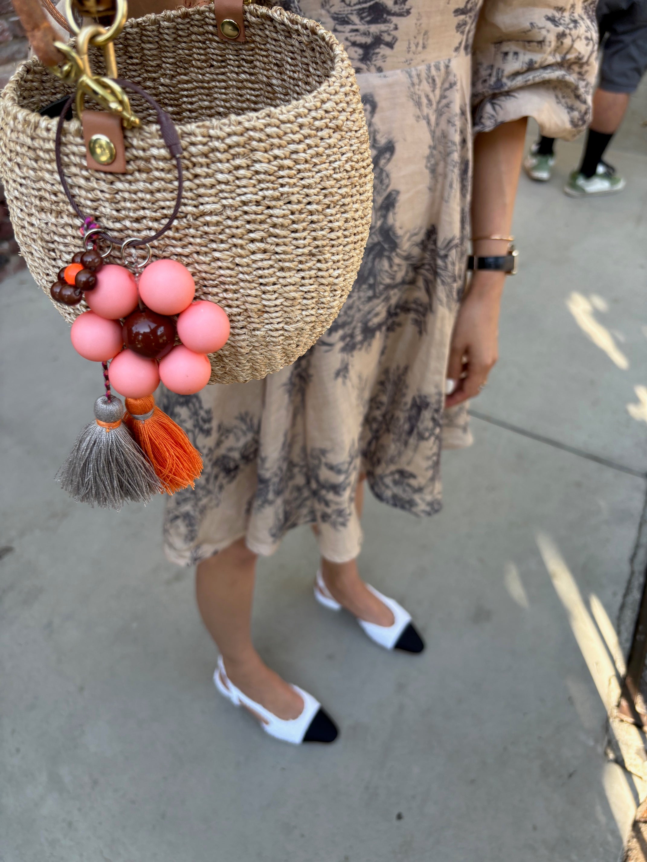 A person is holding a woven straw handbag with a flower keychain attached. The keychain has a peach flower with a brown center and tassels in gray and orange. The person is wearing a beige patterned dress and white shoes with black toe caps.