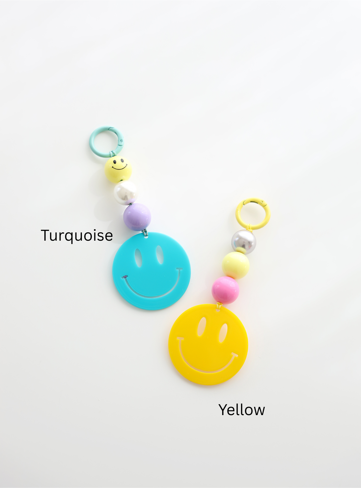 Two colorful smiley face keychains, one in turquoise and one in yellow, each featuring round pastel beads and a large smiley face charm, labeled with their color names on a white background.