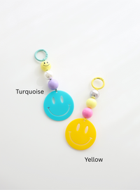 Two colorful smiley face keychains, one in turquoise and one in yellow, each featuring round pastel beads and a large smiley face charm, labeled with their color names on a white background.