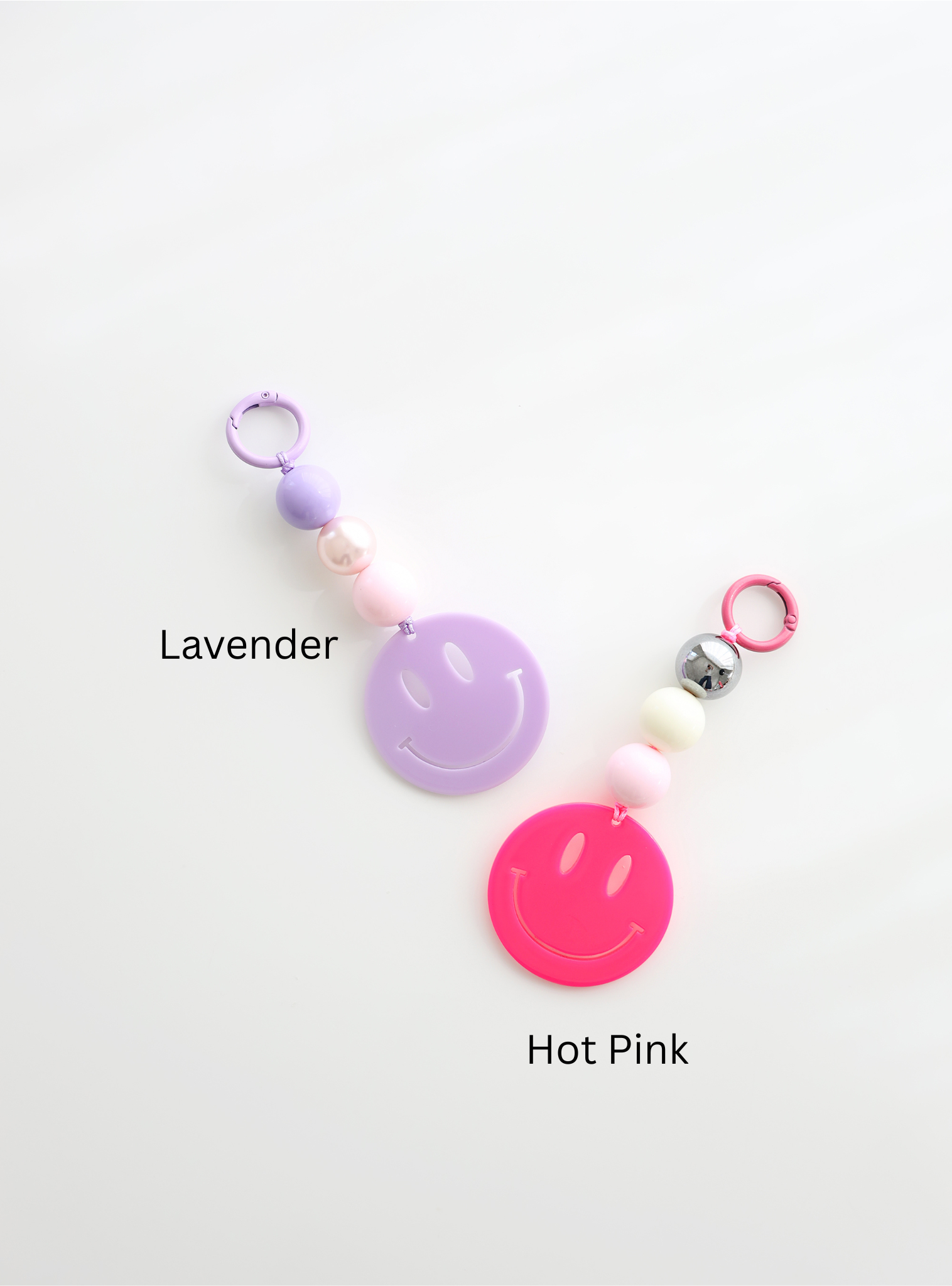 Two colorful smiley face keychains, one in lavender and one in hot pink, each featuring round pastel beads and a large smiley face charm, labeled with their color names on a white background.