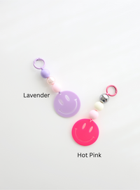 Two colorful smiley face keychains, one in lavender and one in hot pink, each featuring round pastel beads and a large smiley face charm, labeled with their color names on a white background.