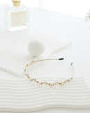 A gold floral headband adorned with small white pearls and crystals, displayed on a wavy white surface with minimalistic props, including a soft white ball and perfume bottle in the background.