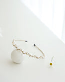 A gold floral headband adorned with small white pearls and crystals, displayed on a white surface with minimalistic props, including a soft white ball.