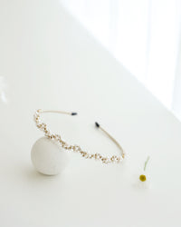 A gold floral headband adorned with small white pearls and crystals, displayed on a white surface with minimalistic props, including a soft white ball.