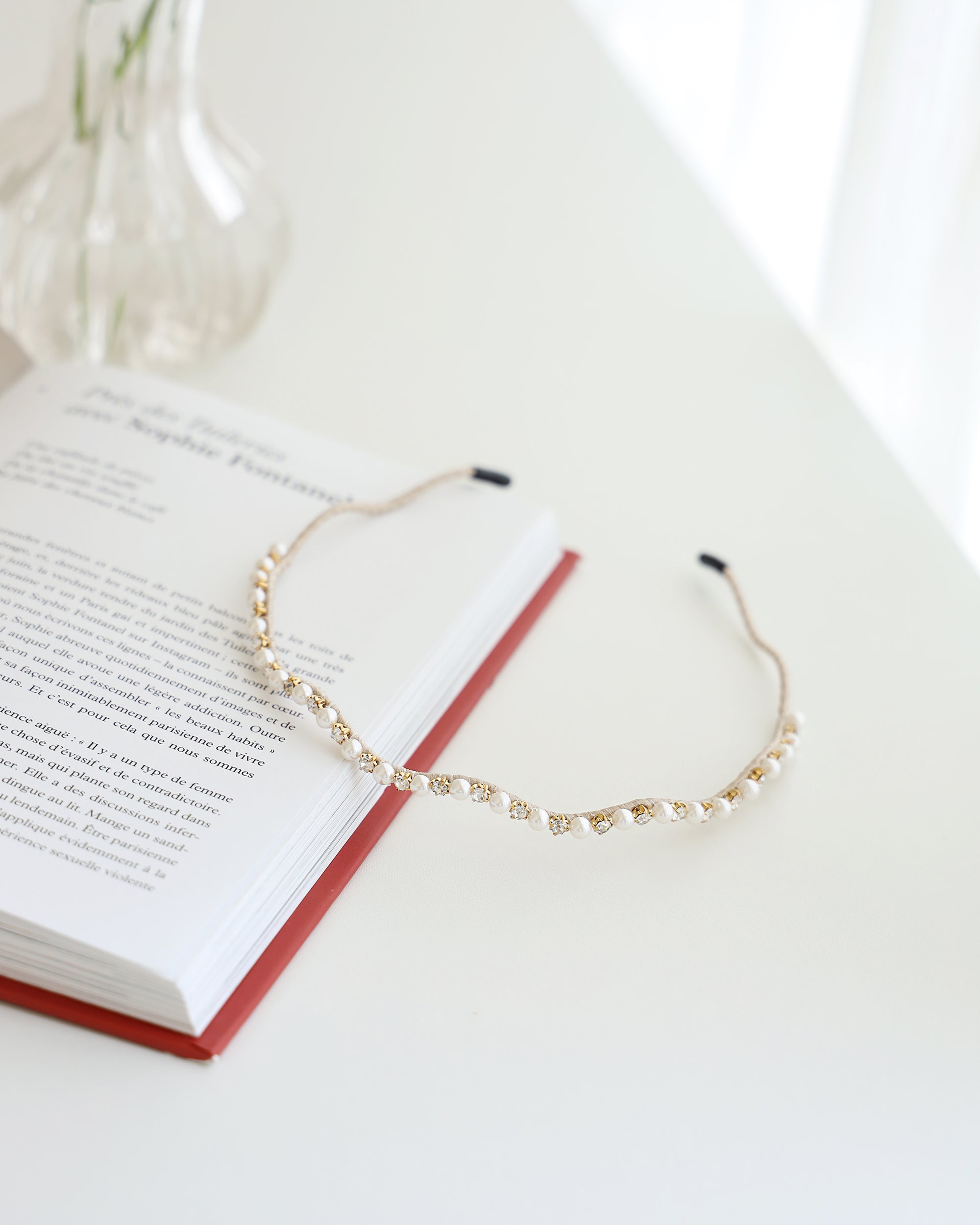A gold vine-style headband with alternating white pearls and crystals, displayed across a white surface with a book and a blurred vase and daisies in the background.