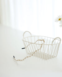 A gold vine-style headband with alternating white pearls and crystals, displayed across a white surface with a white basket and a blurred daisies in the background.