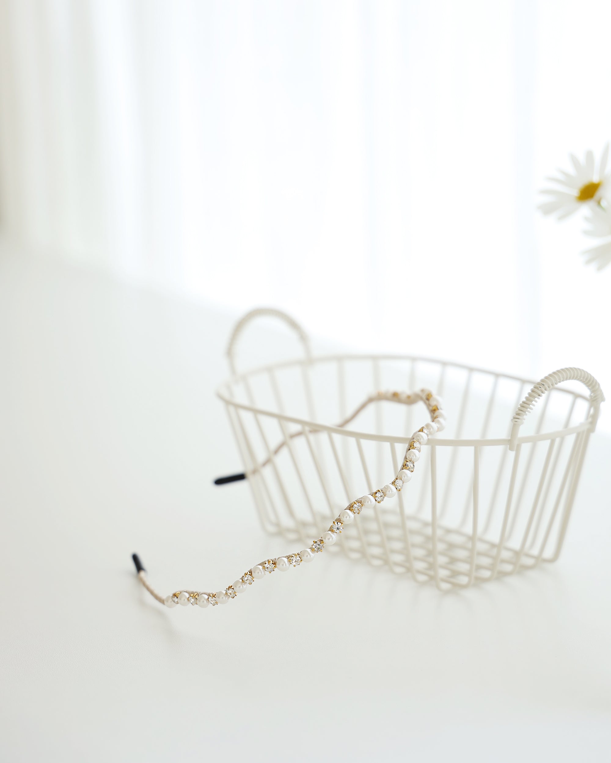 A gold vine-style headband with alternating white pearls and crystals, displayed across a white surface with a white basket and a blurred daisies in the background.