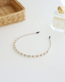 A delicate gold-toned headband featuring small white pearls and sparkling crystals, displayed on white surface with soft natural lighting, perfume bottle and basket.
