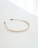 A delicate gold-toned headband featuring small white pearls and sparkling crystals, displayed on white surface with soft natural lighting.