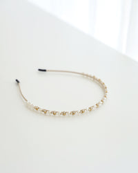 A delicate gold-toned headband featuring small white pearls and sparkling crystals, displayed on white surface with soft natural lighting.