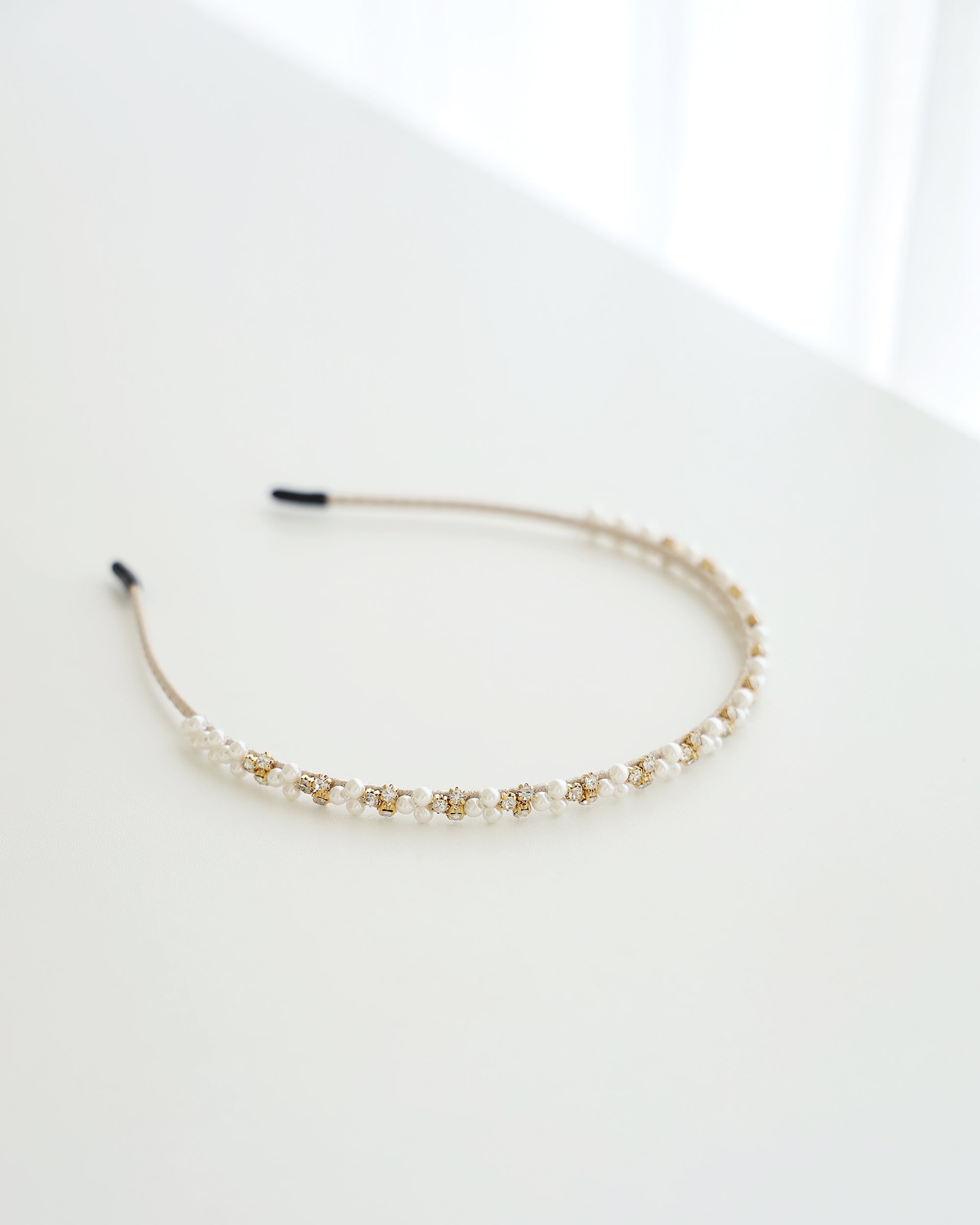 A delicate gold-toned headband featuring small white pearls and sparkling crystals, displayed on white surface with soft natural lighting.