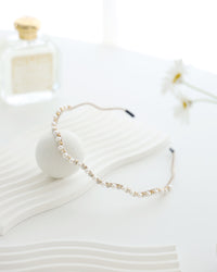 A gold vine-style headband with alternating white pearls and crystals, displayed across a white textured surface with a soft round stone and a blurred perfume bottle and daisies in the background.