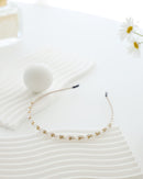 A delicate gold-toned headband featuring small white pearls and sparkling crystals, displayed on a textured white surface with soft natural lighting.