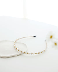 A delicate gold-toned headband featuring small white pearls and sparkling crystals, displayed on white surface with soft natural lighting.