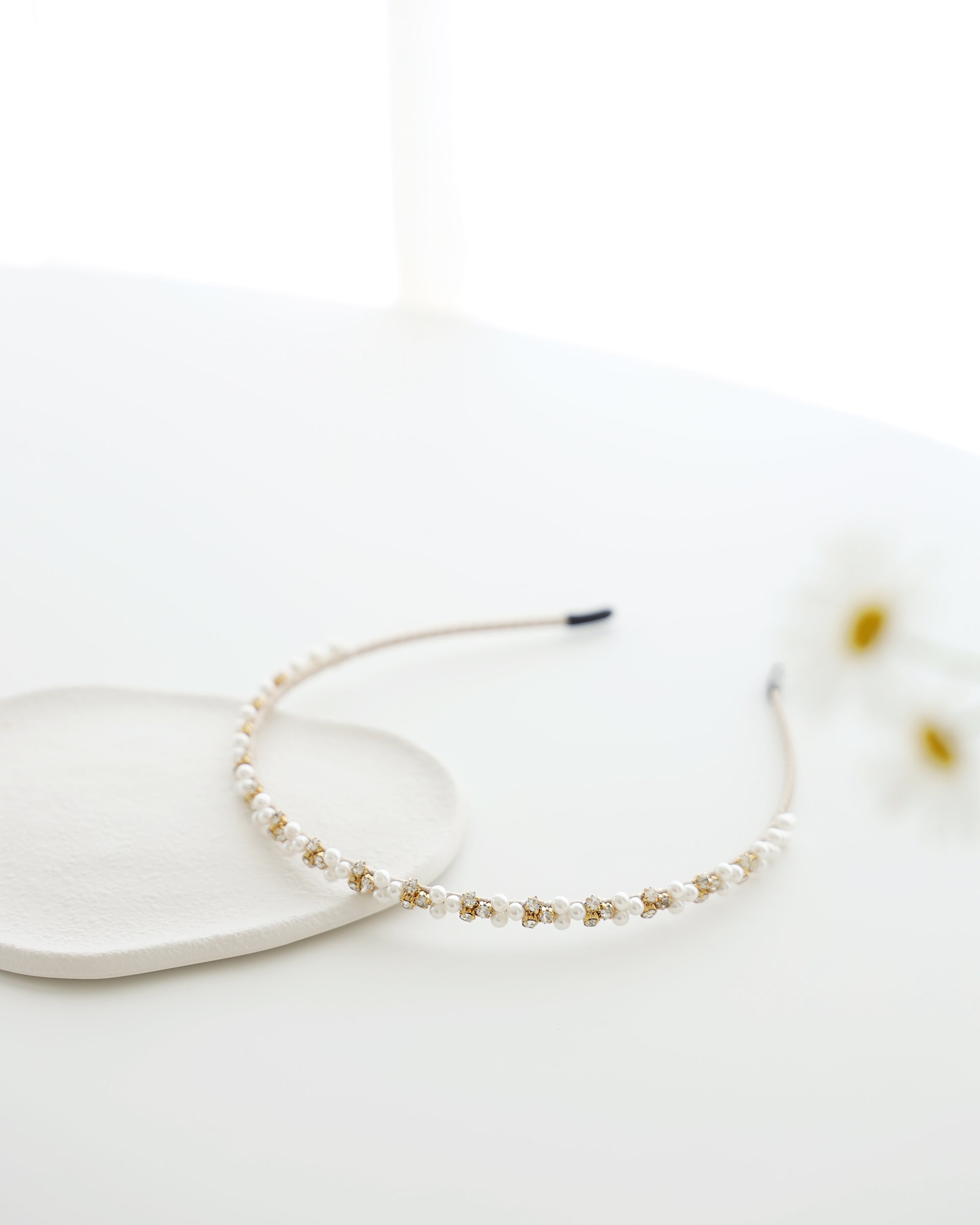 A delicate gold-toned headband featuring small white pearls and sparkling crystals, displayed on white surface with soft natural lighting.