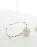 A gold floral headband adorned with small white pearls and crystals, displayed on a white surface with minimalistic props, including a soft white ball.