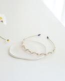 A gold floral headband adorned with small white pearls and crystals, displayed on a white surface with real flower background.