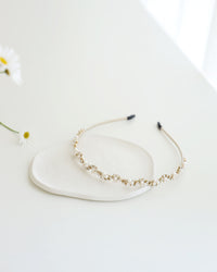 A gold floral headband adorned with small white pearls and crystals, displayed on a white surface with real flower background.