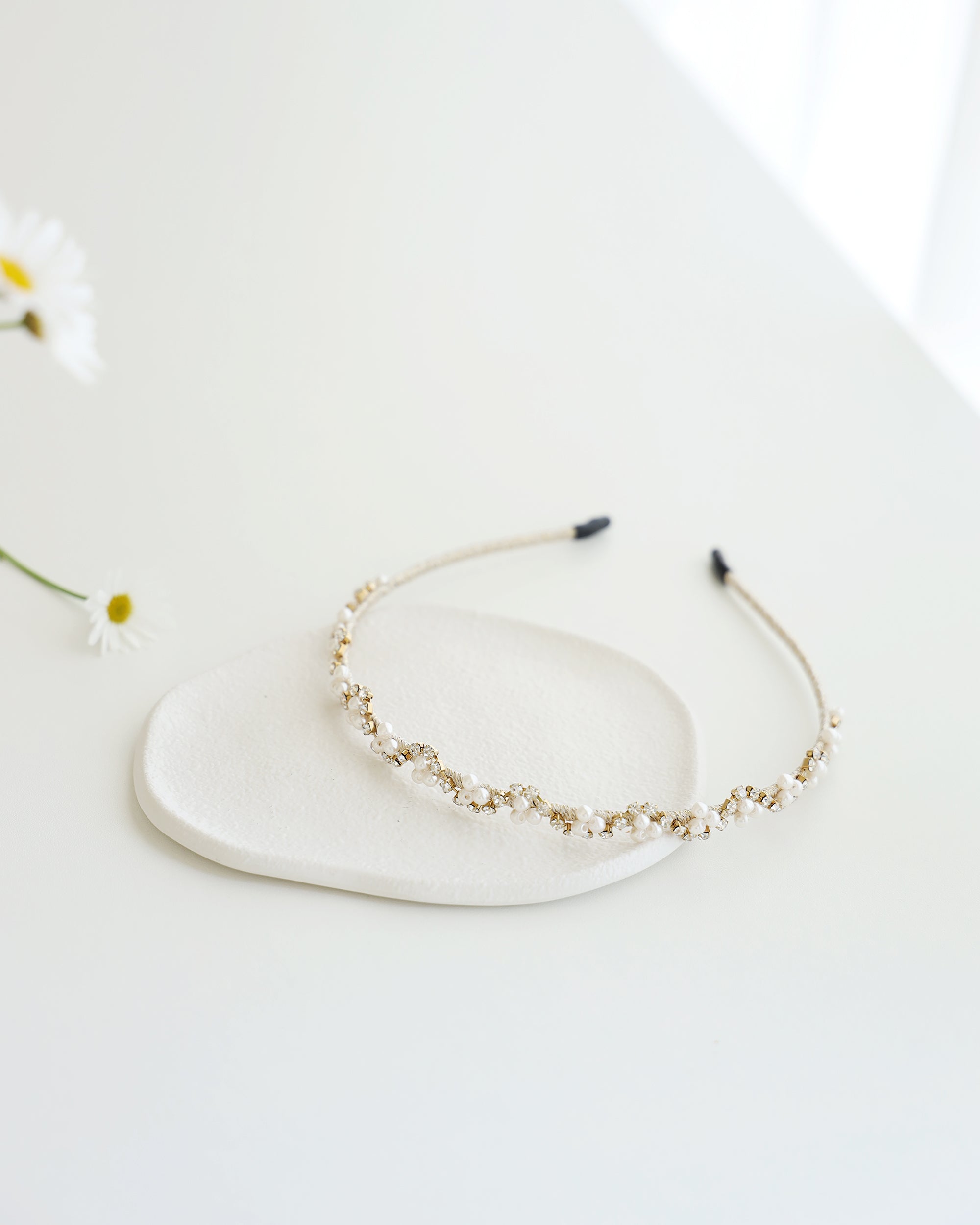 A gold floral headband adorned with small white pearls and crystals, displayed on a white surface with real flower background.
