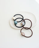 Four elastic hair ties in black and brown, each featuring a glossy mosaic-style bead in ivory, teal, pastel, and mauve tones, displayed on a textured white dish.