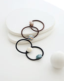 Four elastic hair ties in black and brown, each featuring a glossy mosaic-style bead in ivory, teal, pastel, and mauve tones, displayed on a textured white dish.