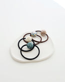 Four elastic hair ties in black and brown, each featuring a glossy mosaic-style bead in ivory, teal, pastel, and mauve tones, displayed on a textured white dish.