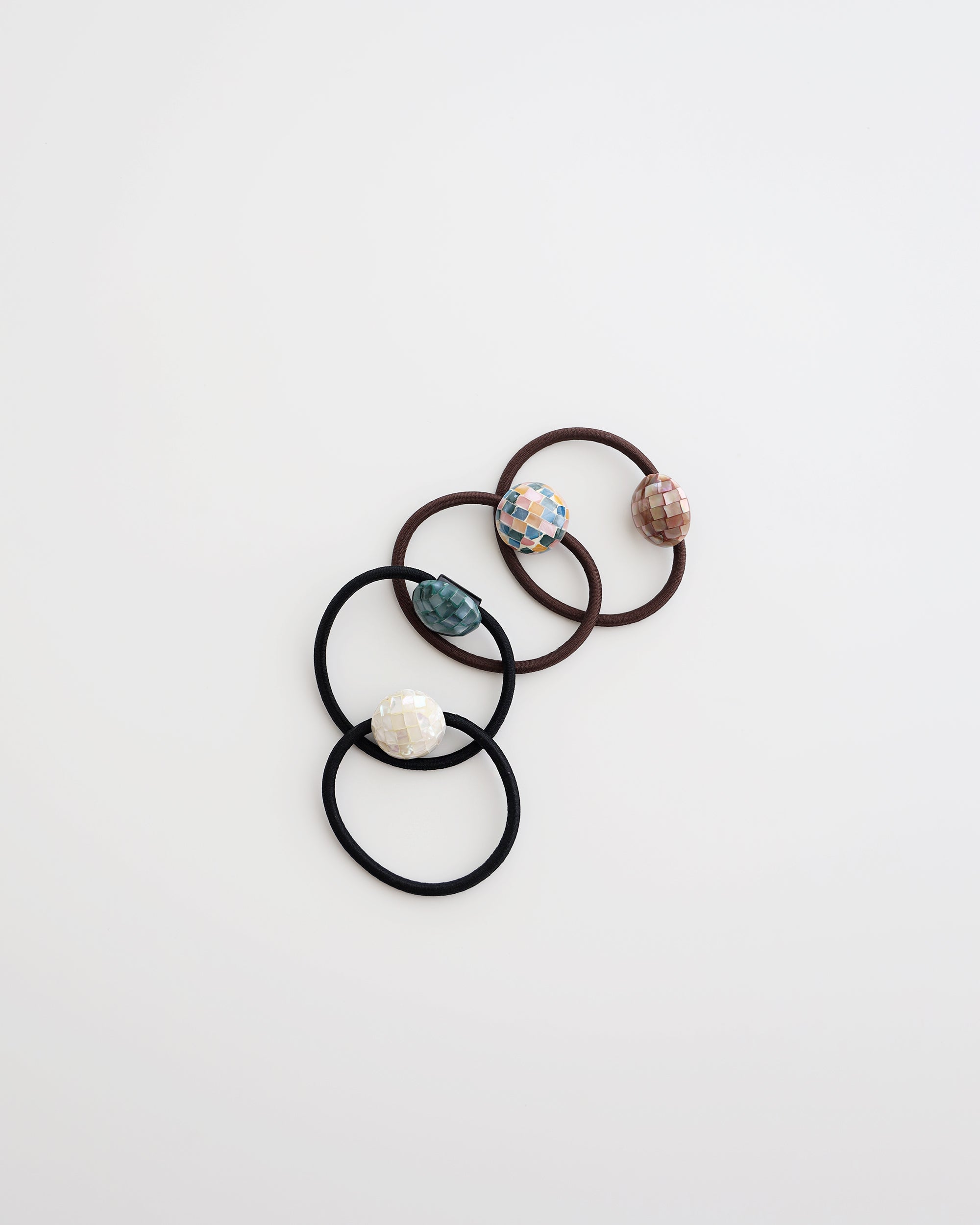 Four elastic hair ties in black and brown, each featuring a glossy mosaic-style bead in ivory, teal, pastel, and mauve tones, displayed on white background.