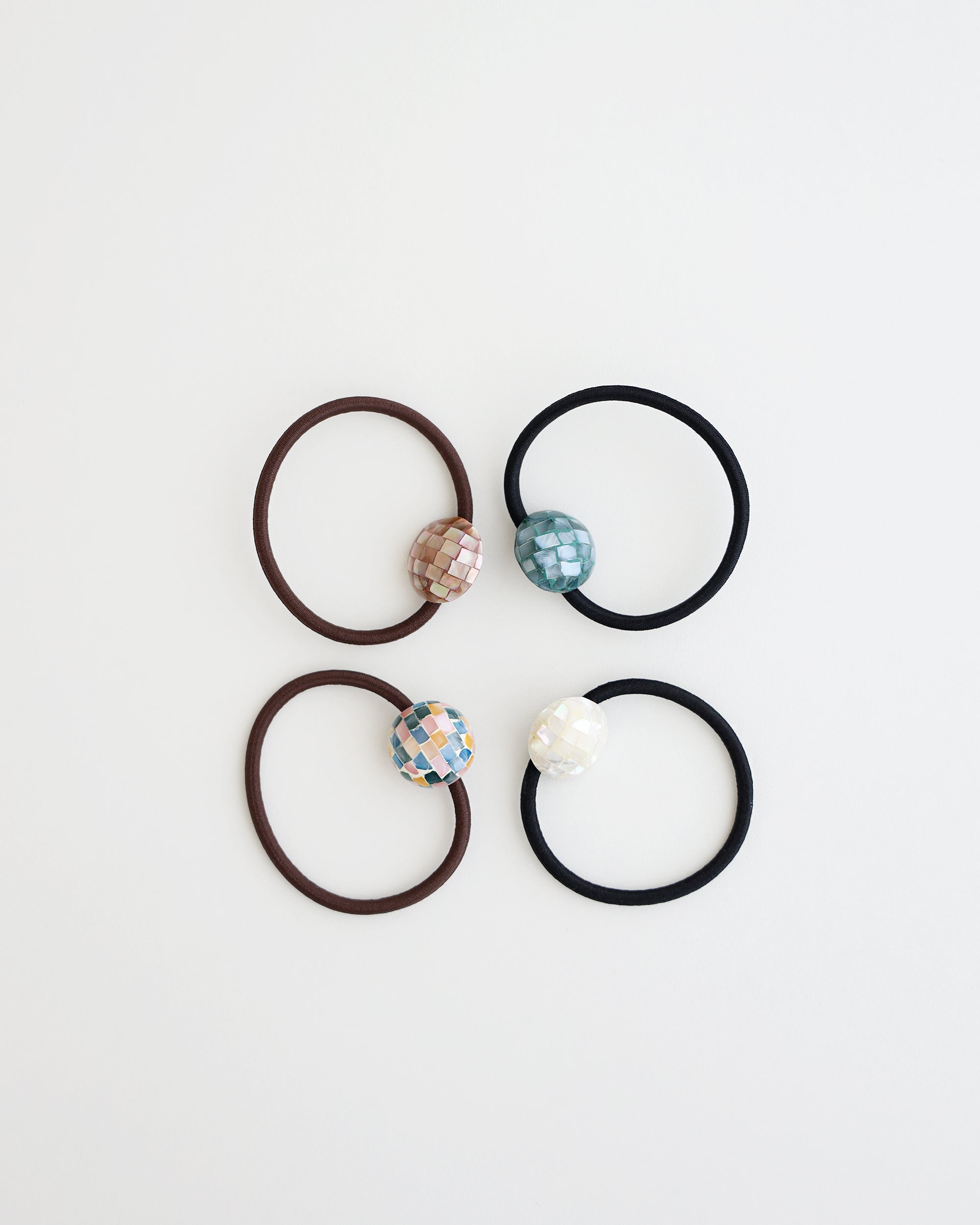 Four elastic hair ties in black and brown, each featuring a glossy mosaic-style bead in ivory, teal, pastel, and mauve tones, displayed on white background.