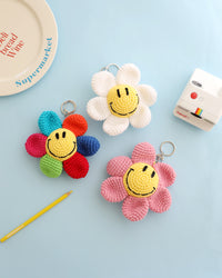 Three crocheted flower keychains with cheerful yellow smiley face centers, shown in white, pink, and multicolor petals, displayed on a light blue background with playful props like a Polaroid camera, a yellow pencil, and a decorative plate.