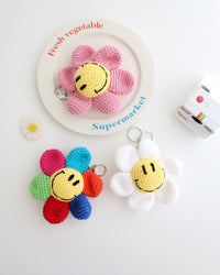 Three crocheted flower keychains with cheerful yellow smiley face centers, shown in white, pink, and multicolor petals, displayed on a white background with playful props like a Polaroid camera, a decorative daisy flower.