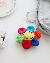A multicolor crocheted flower keychain with a bright yellow smiley face center is shown clipped to the zipper of a light gray bag. The petals are pink, green, red, blue, turquoise, and orange, creating a cheerful rainbow effect.