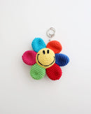 A multicolor crocheted flower keychain with a bright yellow smiley face center is shown on a white background. The petals are pink, green, red, blue, turquoise, and orange, creating a cheerful rainbow effect.