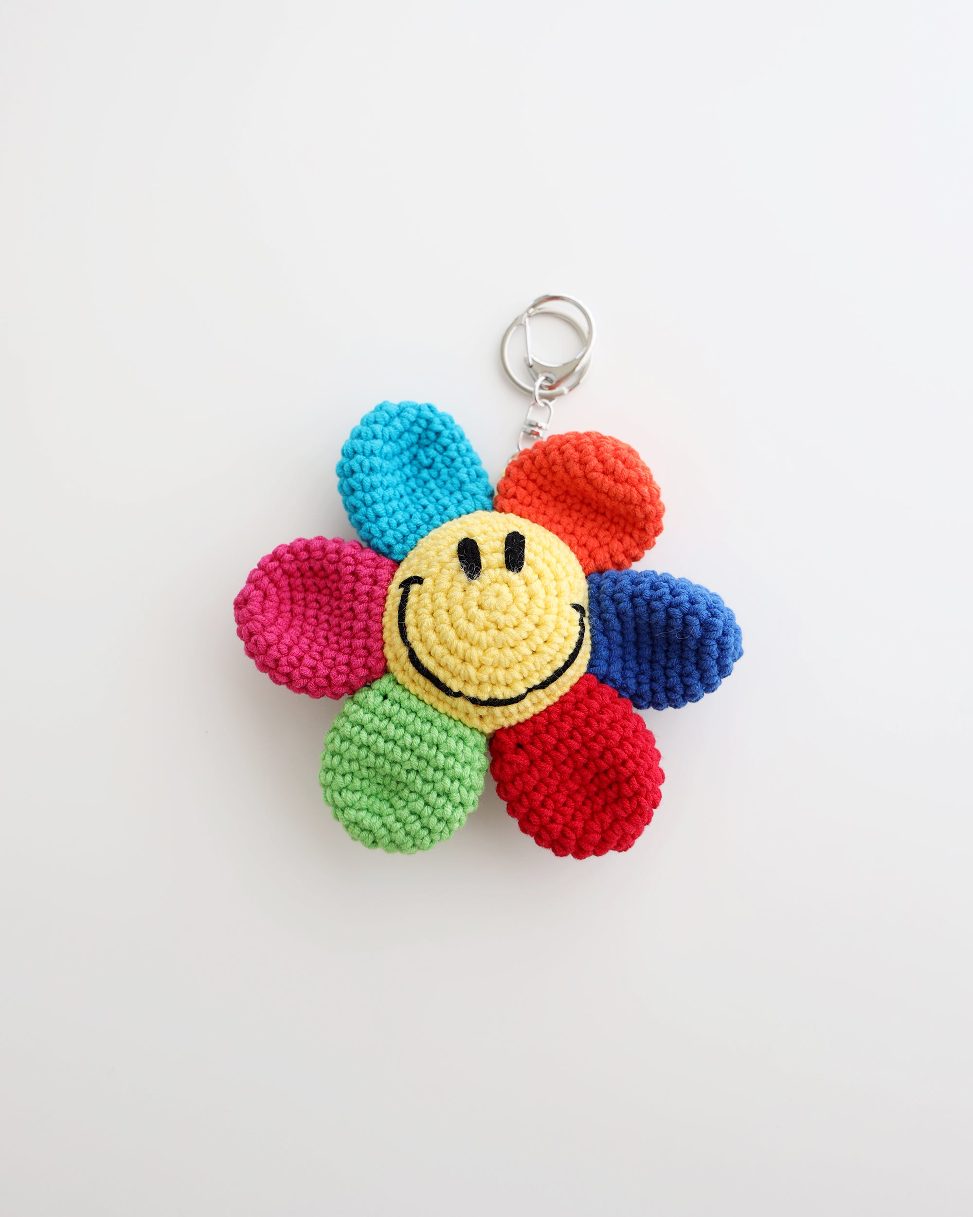 A multicolor crocheted flower keychain with a bright yellow smiley face center is shown on a white background. The petals are pink, green, red, blue, turquoise, and orange, creating a cheerful rainbow effect.