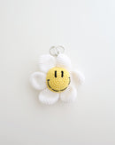 A crocheted flower keychain with soft white petals and a bright yellow smiley face center is displayed. It features a silver keyring at the top for easy attachment to bags, keys, or pouches.