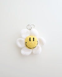 A crocheted flower keychain with soft white petals and a bright yellow smiley face center is displayed. It features a silver keyring at the top for easy attachment to bags, keys, or pouches.