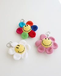 Three crocheted flower keychains with cheerful yellow smiley face centers, shown in white, pink, and rainbow petals, displayed on a white background.