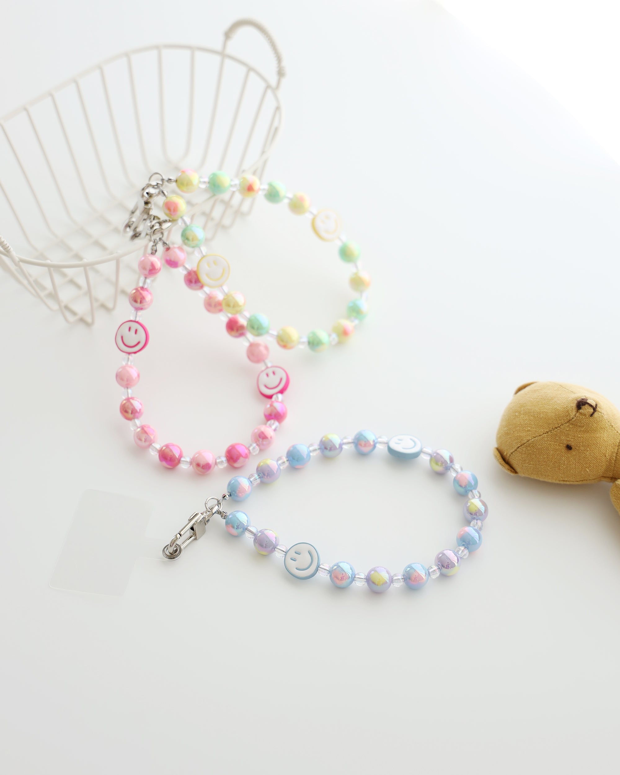 Three pastel beaded phone straps with glossy round beads and one smiley face bead. Available in pink, blue, or yellow color themes. Comes with a clear plastic phone tab that fits between the phone and case, with a metal clasp to attach the strap displayed on white background