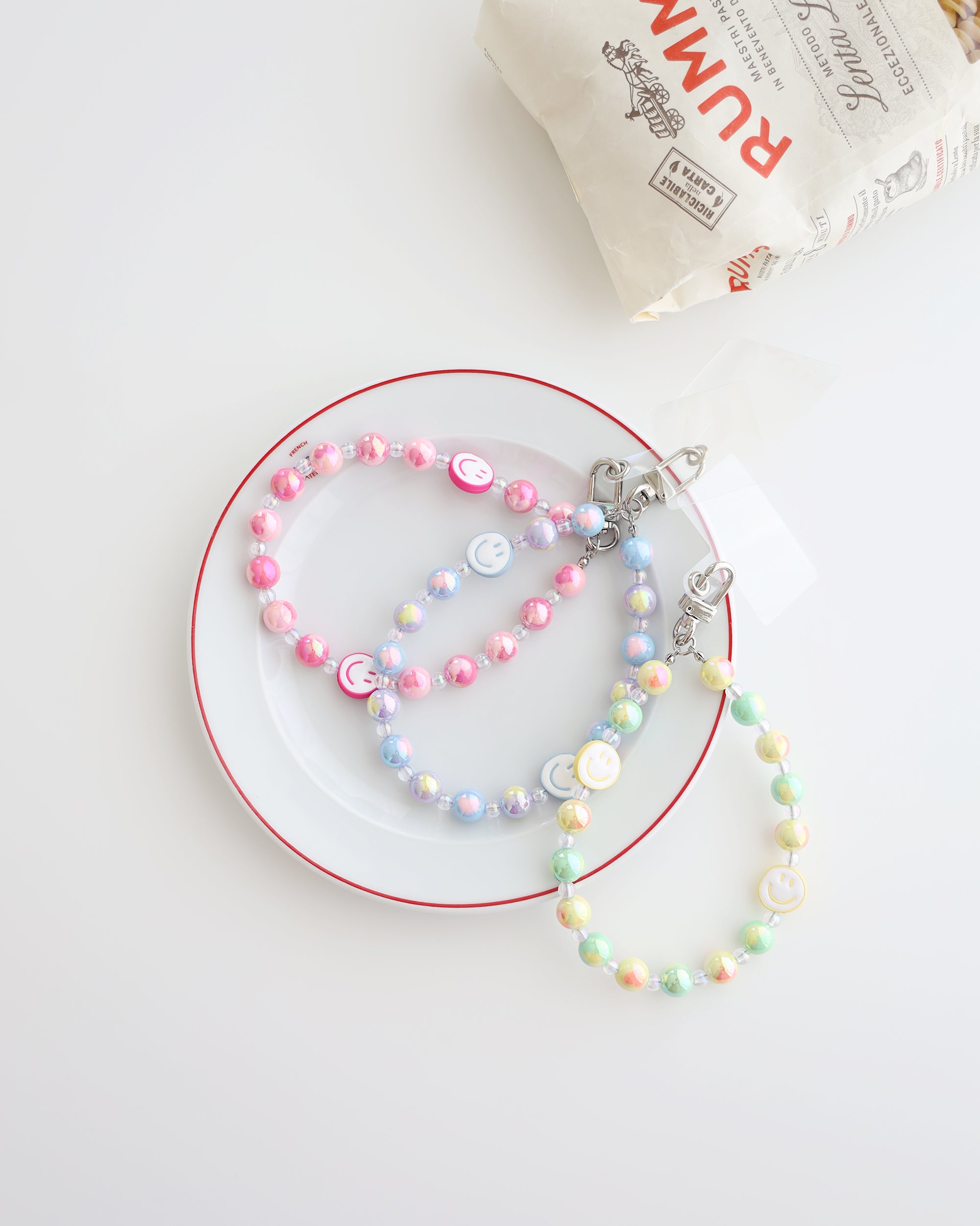 Three Pastel beaded phone straps with glossy round beads and one smiley face bead. Available in pink, blue, or yellow color themes. Comes with a clear plastic phone tab that fits between the phone and case, with a metal clasp to attach the strap displayed on a white plate with white background and one bag of pasta 
