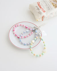 Three Pastel beaded phone straps with glossy round beads and one smiley face bead. Available in pink, blue, or yellow color themes. Comes with a clear plastic phone tab that fits between the phone and case, with a metal clasp to attach the strap displayed on a white plate with white background and one bag of pasta 