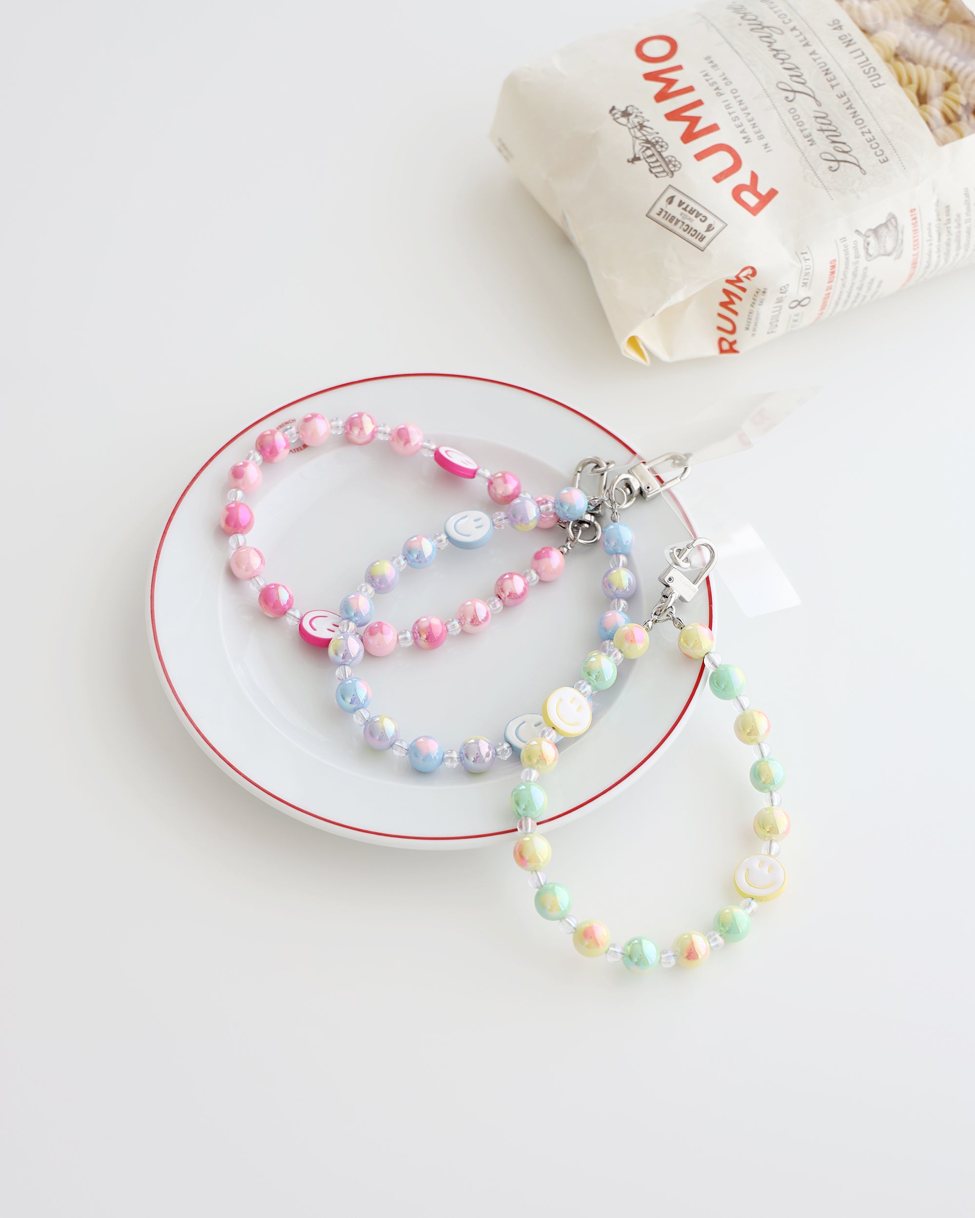 Three Pastel beaded phone straps with glossy round beads and one smiley face bead. Available in pink, blue, or yellow color themes. Comes with a clear plastic phone tab that fits between the phone and case, with a metal clasp to attach the strap displayed on a white plate with white background and one bag of pasta 