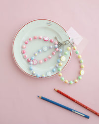 Pastel beaded phone strap with glossy round beads and one smiley face bead. Available in pink, blue, or yellow color themes. Comes with a clear plastic phone tab that fits between the phone and case, with a metal clasp to attach the strap displayed on a white plate with pink background and two pencils