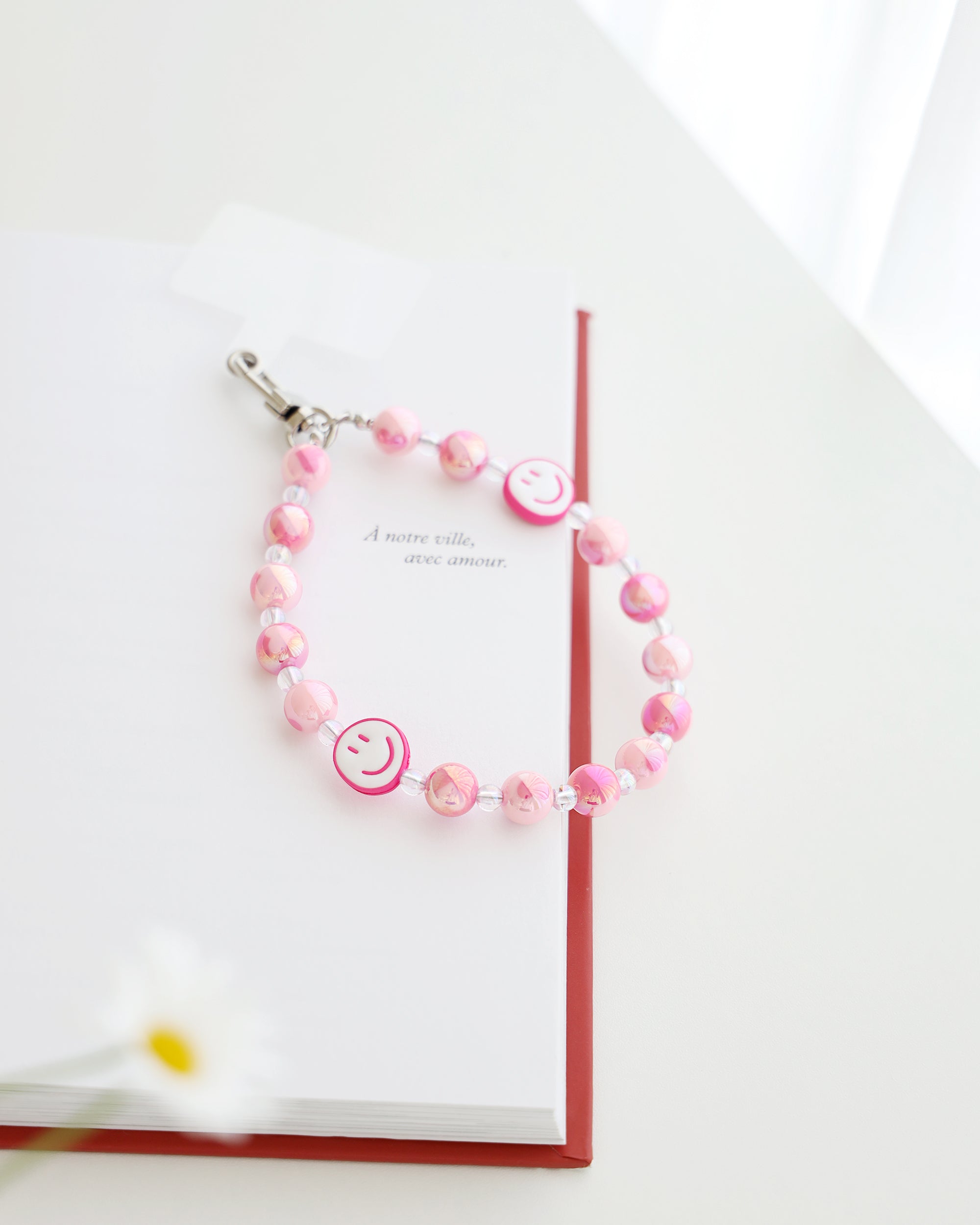 Pastel pink beaded phone strap with glossy round beads and one white smiley face bead with pink smile. Comes with a clear plastic phone tab and metal clasp for attachment displayed on a book.