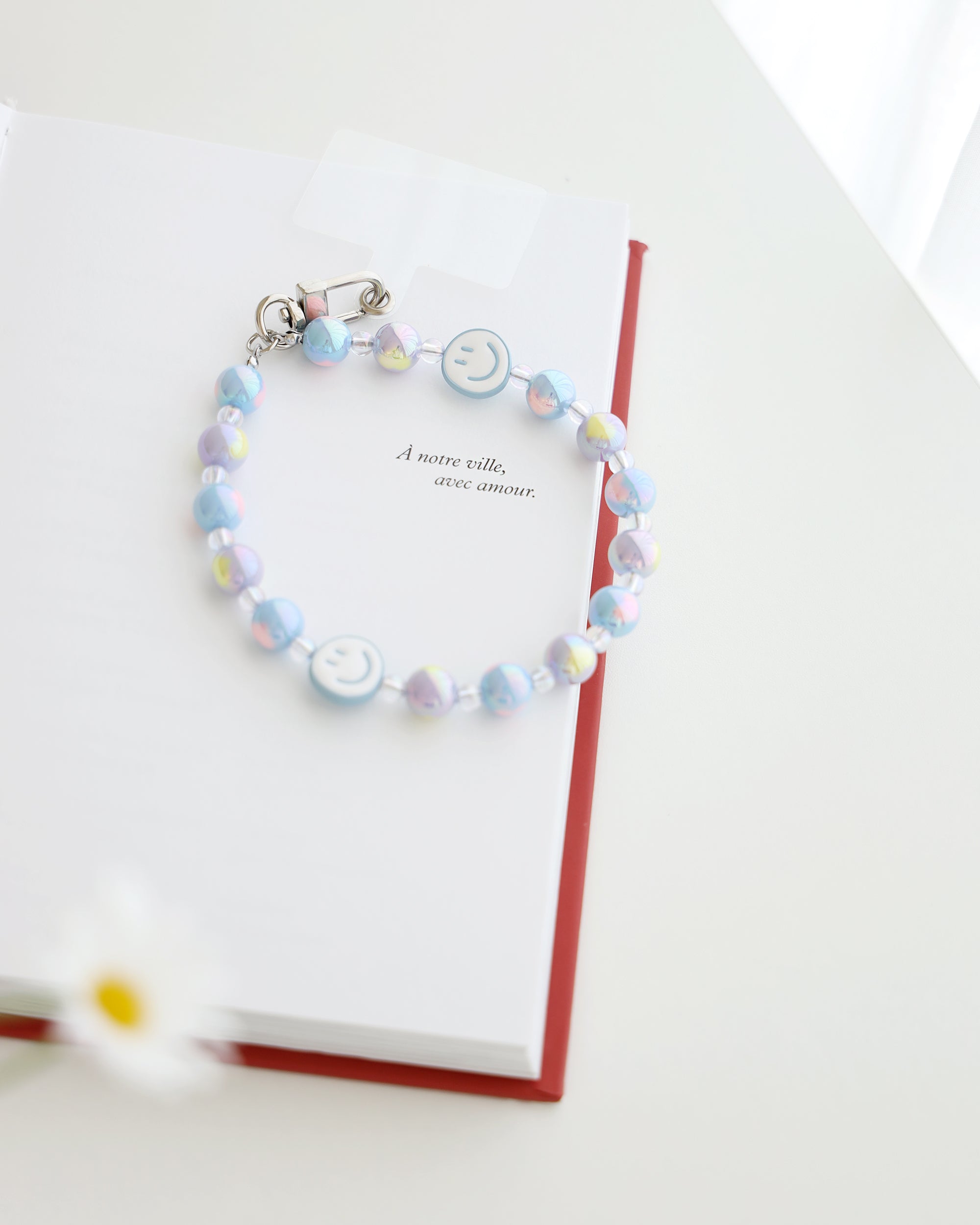 Pastel blue and lavender beaded phone strap with glossy round beads and one white smiley face bead with blue smile. Comes with a clear plastic phone tab and metal clasp for attachment displayed on a book