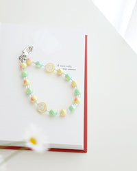 Pastel blue and lavender beaded phone strap with glossy round beads and one white smiley face bead with blue smile. Comes with a clear plastic phone tab and metal clasp for attachment displayed on a book