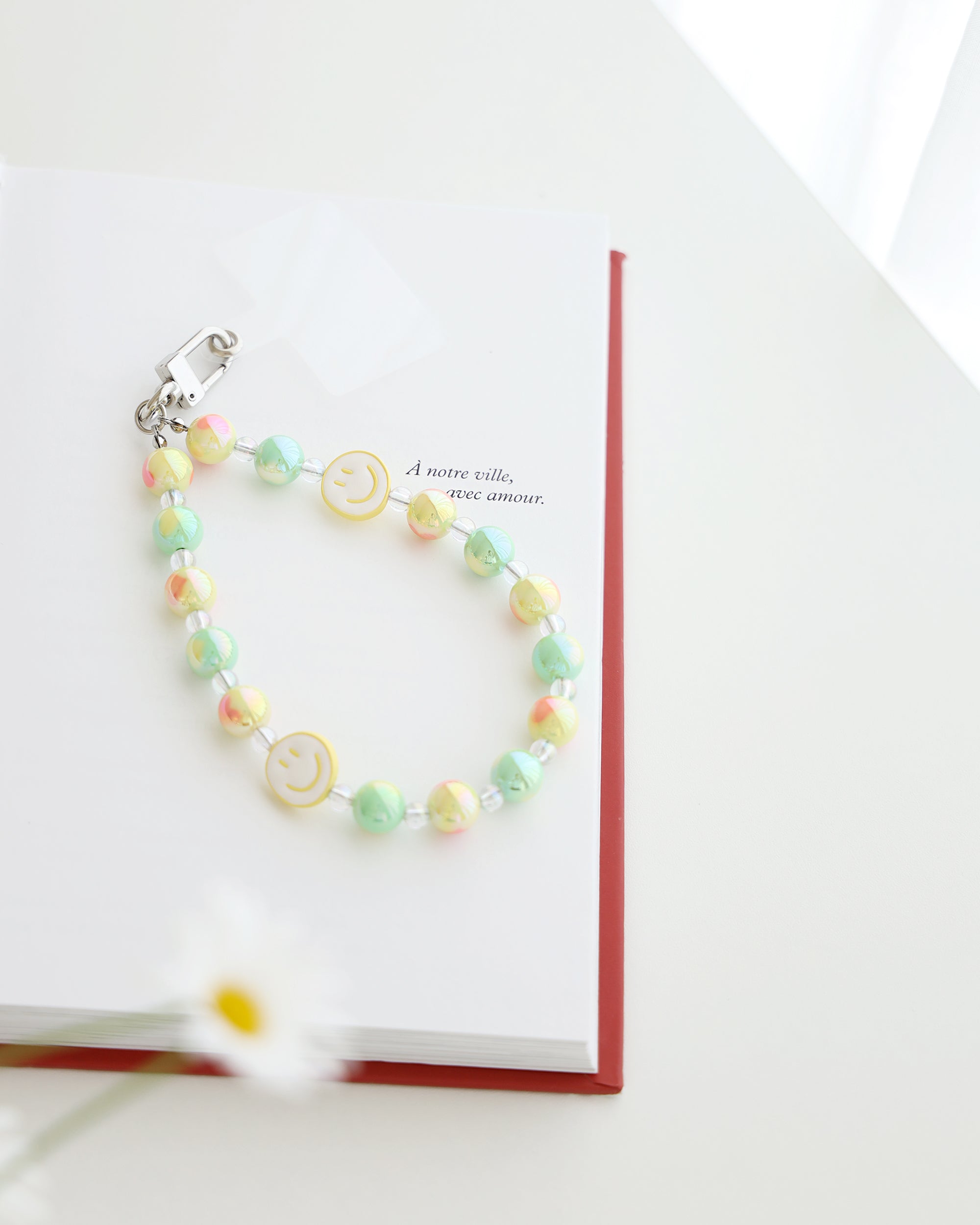 Pastel blue and lavender beaded phone strap with glossy round beads and one white smiley face bead with blue smile. Comes with a clear plastic phone tab and metal clasp for attachment displayed on a book
