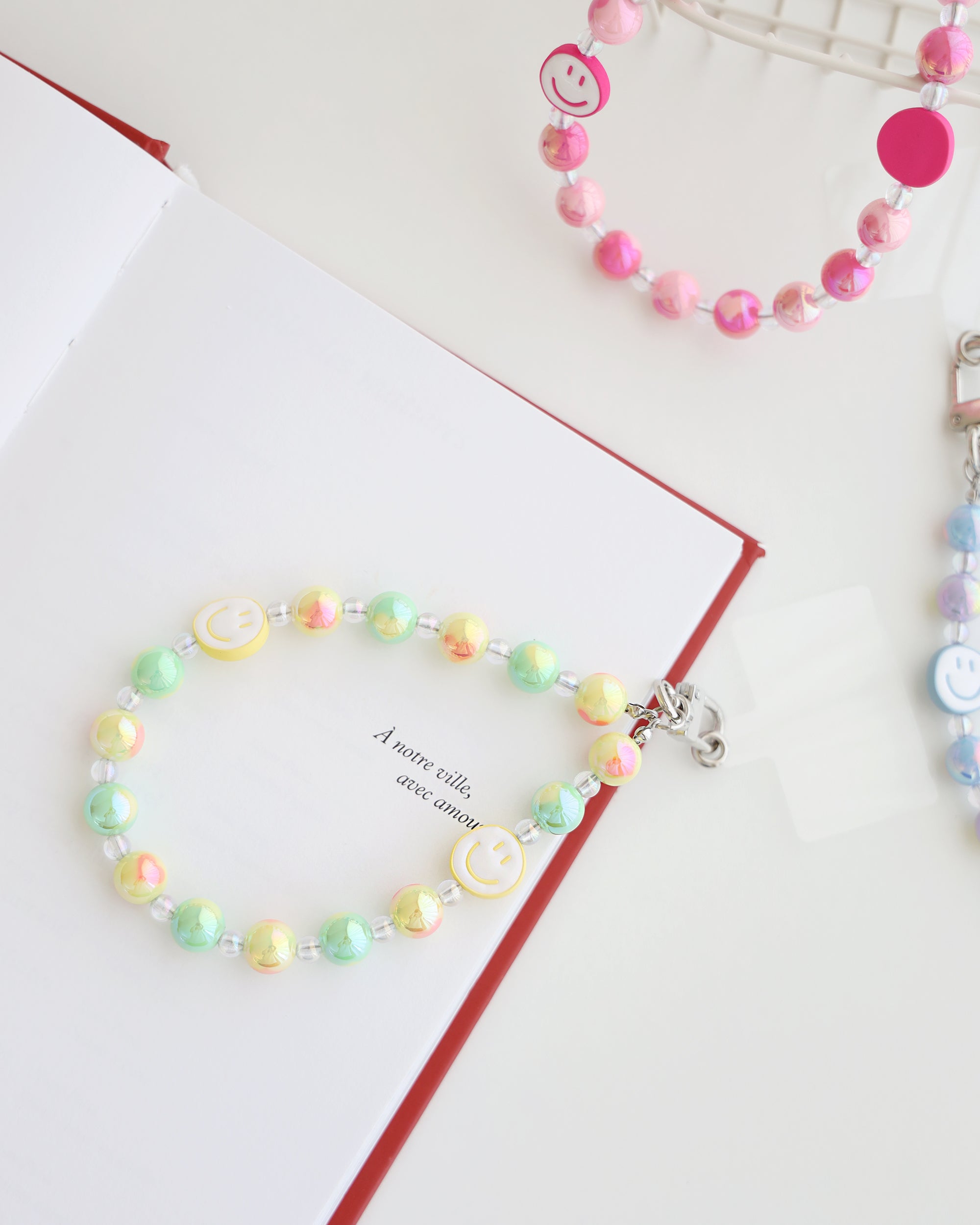 Pastel yellow, green, and peach beaded phone strap with glossy round beads and two white smiley face beads with yellow smiles. Attached to a clear plastic phone tab with a metal clasp. Pink and blue color variants are partially visible in the background.