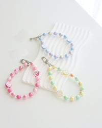 Three pastel beaded phone straps with glossy round beads and two smiley face beads each. One strap is pink with white smiley faces and pink smiles, one is blue and lavender with white smiley faces and blue smiles, and one is yellow, green, and peach with white smiley faces and yellow smiles. Each strap is attached to a clear plastic phone tab with a metal clasp.