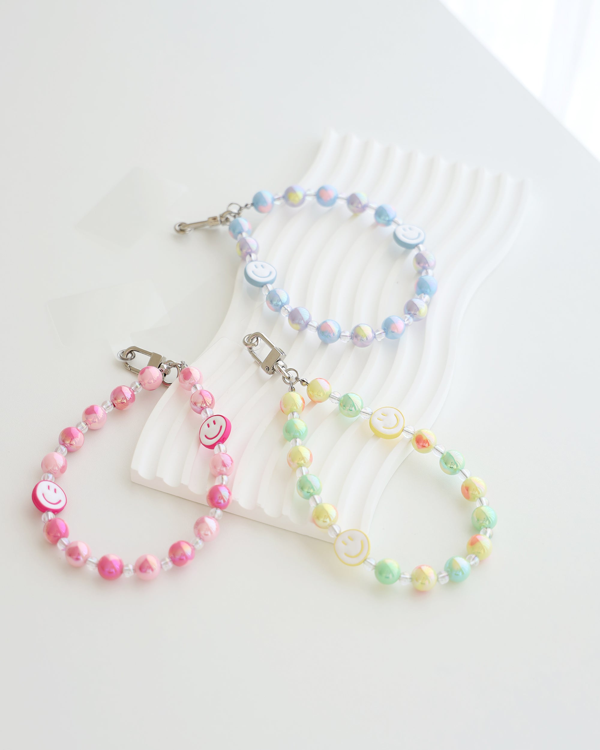 Three pastel beaded phone straps with glossy round beads and two smiley face beads each. One strap is pink with white smiley faces and pink smiles, one is blue and lavender with white smiley faces and blue smiles, and one is yellow, green, and peach with white smiley faces and yellow smiles. Each strap is attached to a clear plastic phone tab with a metal clasp.