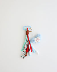 A keychain charm made with multiple knotted cord tassels and a sky blue crocheted heart which includes a sky blue metal carabiner clip for attaching to bags, keys, or other items. The tassels are made of multi-color cords, and the crochet heart features a stitched label.