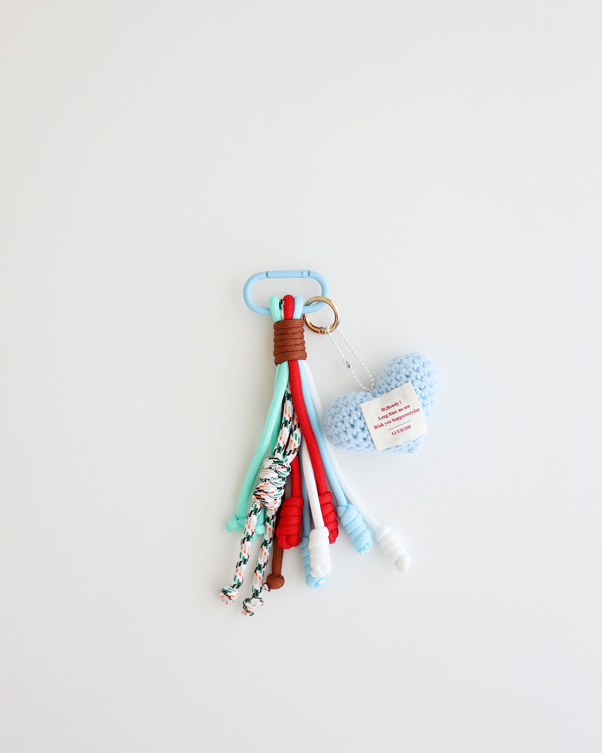 A keychain charm made with multiple knotted cord tassels and a sky blue crocheted heart which includes a sky blue metal carabiner clip for attaching to bags, keys, or other items. The tassels are made of multi-color cords, and the crochet heart features a stitched label.